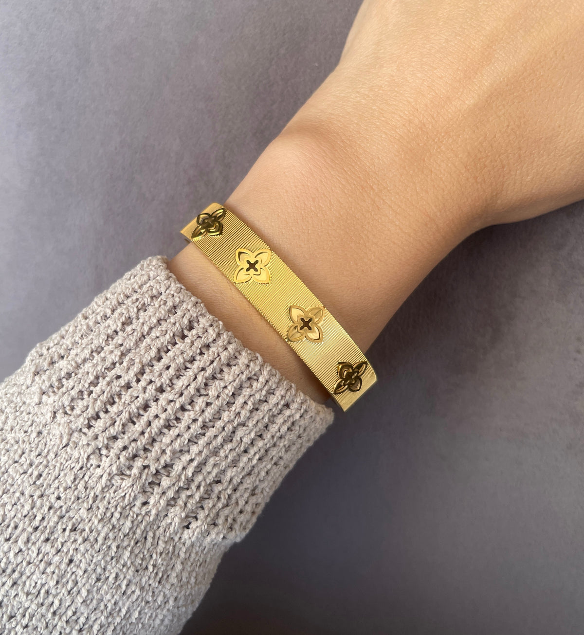 Gold textured floral clover bangle bracelet worn on model wrist with beige knit sweater.
