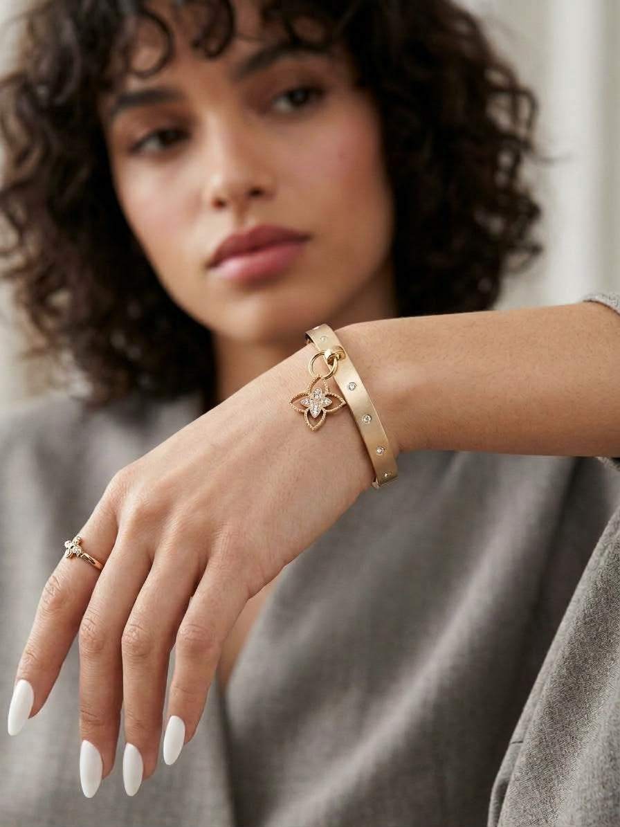 Gold textured bangle bracelet with cz accents and dangling floral clover charm on model wrist.
