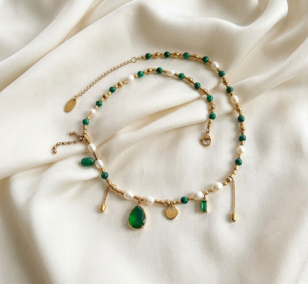 A beaded charm necklace with an extension chain resting on a draped, off-white satin surface. The necklace combines white pearls, green beads, and gold spacers, adorned with various charms including a prominent teardrop-shaped green gemstone pendant at the center, a gold heart charm, a rectangular green jewel, and dangling gold chains.
