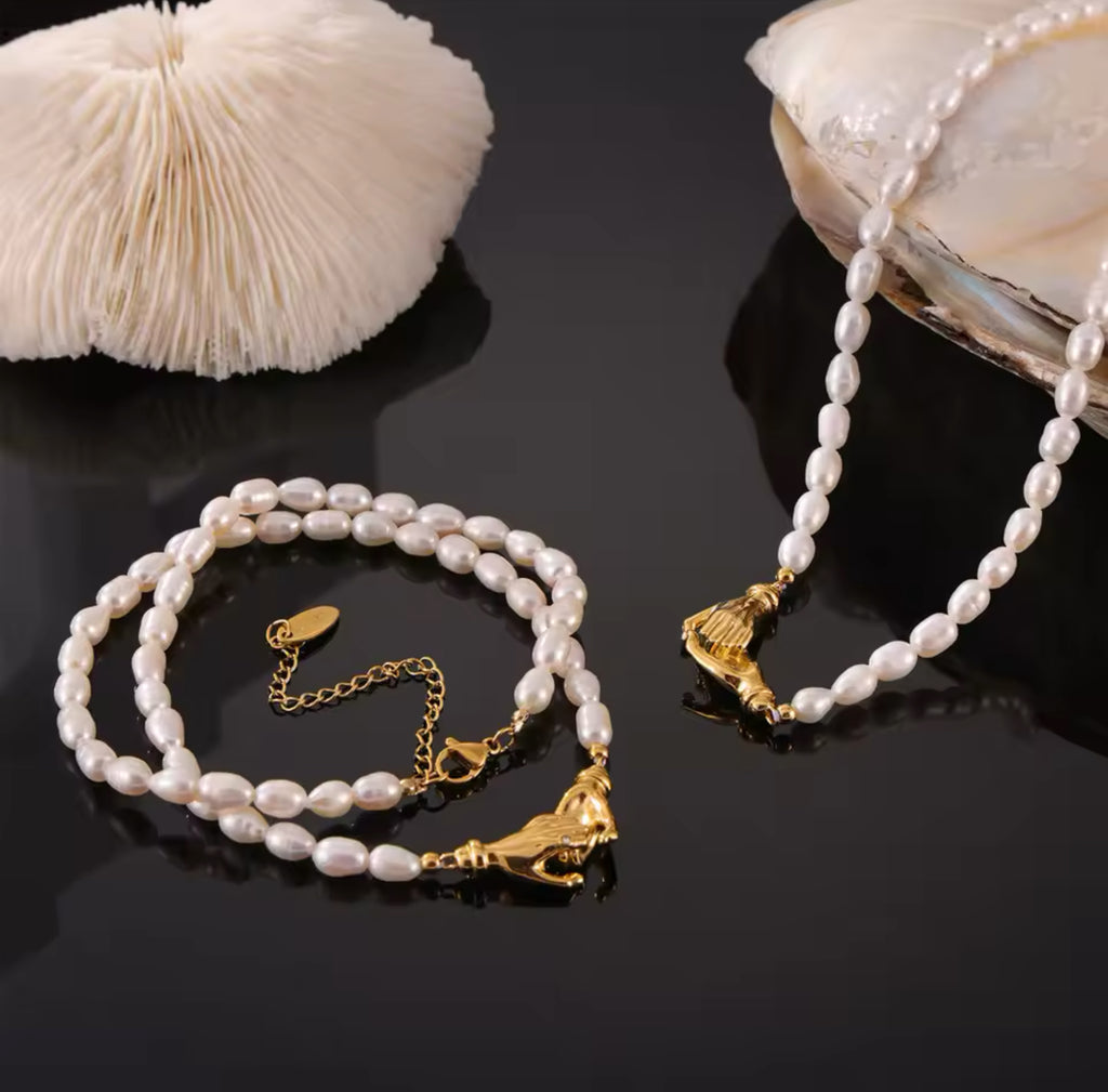 White freshwater pearl necklaces featuring intricate gold interlocking hand clasps, styled on a reflective dark surface with a white coral accent and seashell background.