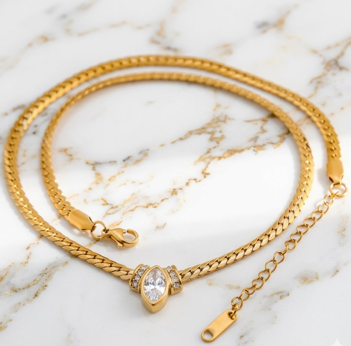 A gold curb-style chain necklace with a central marquise-cut diamond pendant resting on a light-colored marble surface. The necklace includes a lobster claw clasp and an adjustable extender chain at the ends.
