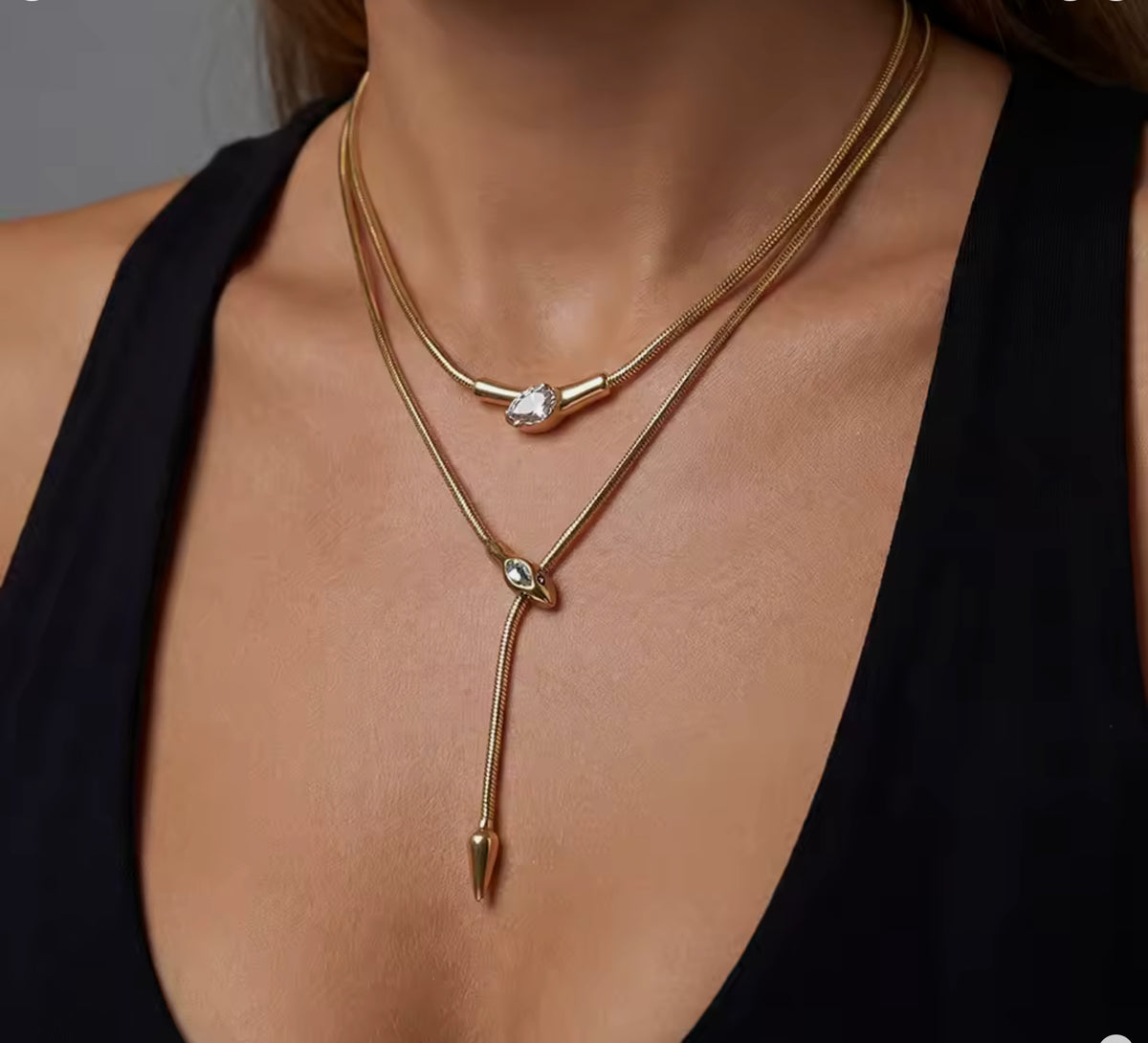 Close-up of a model wearing layered gold snake-chain necklaces: a shorter collar with a teardrop charm and a longer lariat-style necklace featuring a serpent head sliding pendant with a pointed tail drop, set against a black V-neck top.
