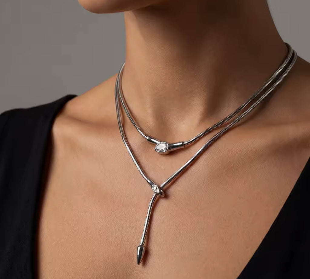 Close-up of a model wearing layered silver snake-chain necklaces: a short collar with a single bezel-set round stone centerpiece and a longer lariat necklace with a serpent head clasp and a pointed drop tail, styled against a black top.