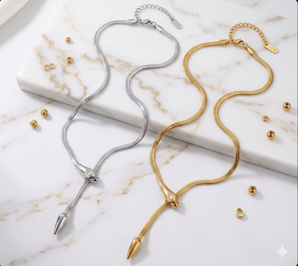 Silver and gold snake-chain lariat necklaces with serpent head clasps and pointed drop tails, displayed on a marble surface with loose golden beads.