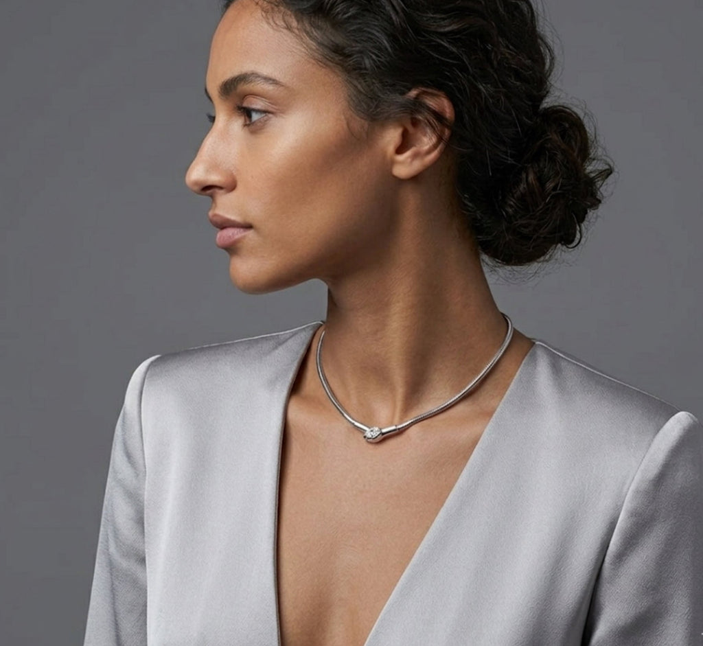 Side-profile portrait of a woman with dark hair in a low bun wearing a silver rigid collar necklace with a pave-set diamond snake head center, paired with a silver-grey plunging V-neck jacket.