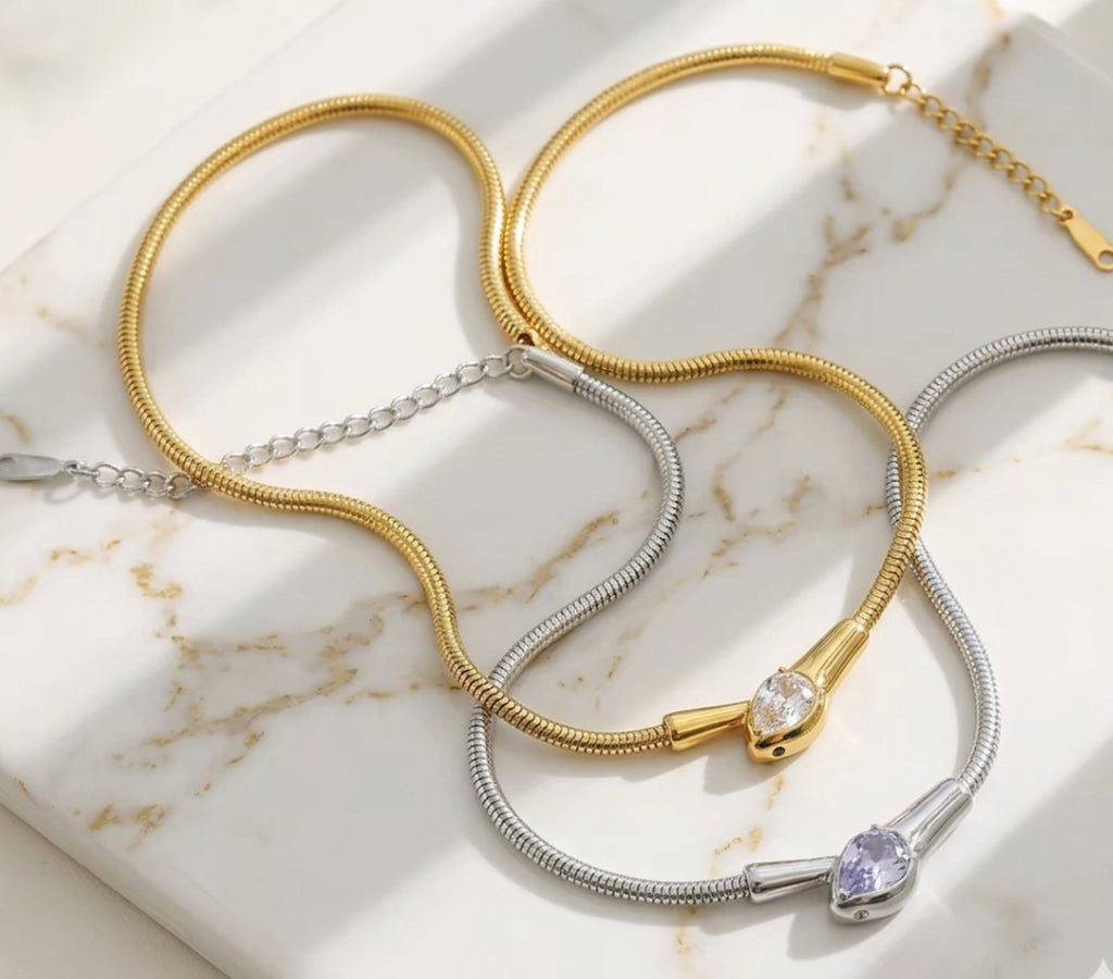 Gold and silver snake-chain collar necklaces with pave-set serpent head centerpieces, arranged on a light-colored marble surface.