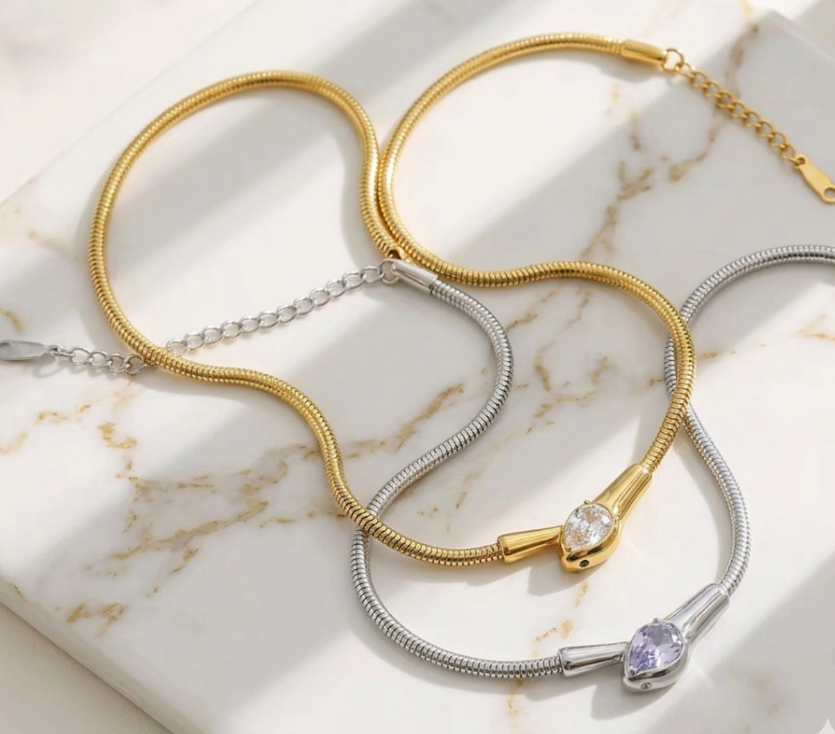 Gold and silver snake-chain collar necklaces with pave-set serpent head centerpieces, arranged on a light-colored marble surface.