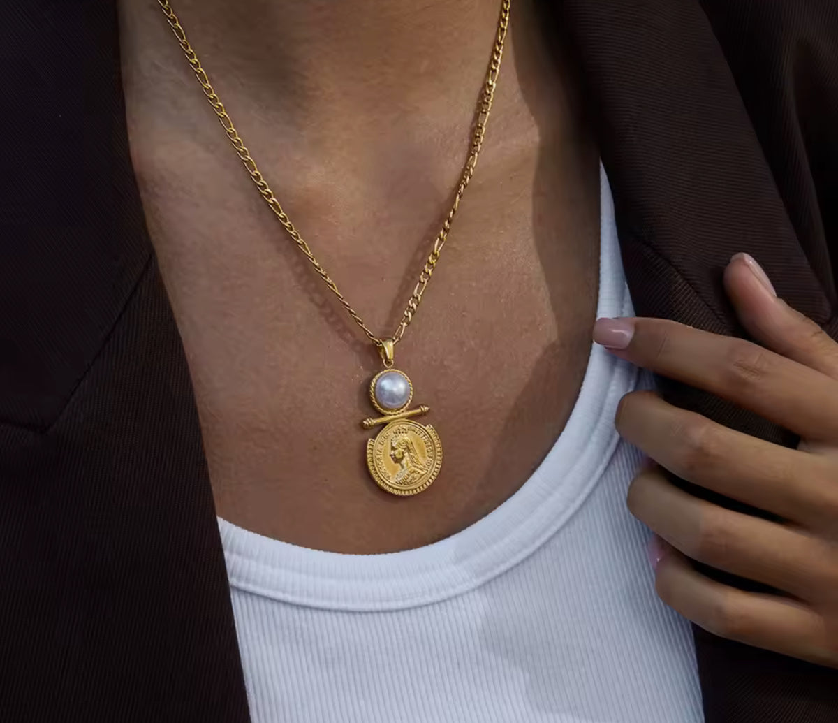 A close-up of a person wearing a gold pendant necklace with a round coin-style charm and a white pearl accent, styled over a white ribbed top and dark blazer.