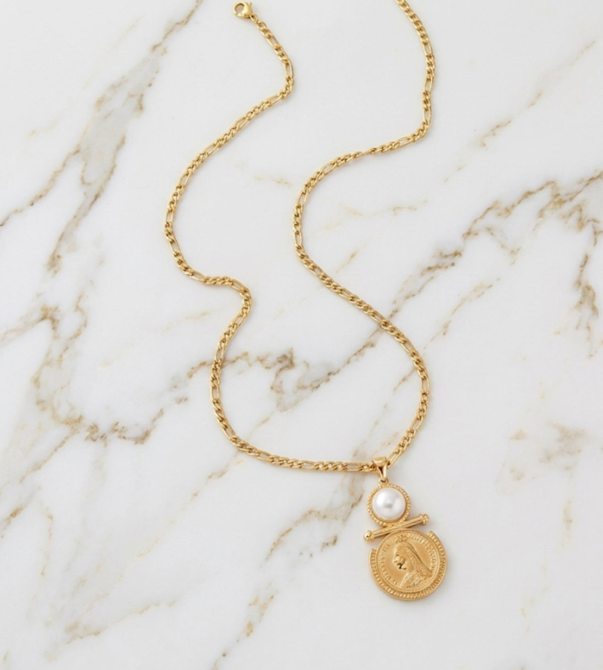 A gold chain necklace with a round gold medallion pendant and a small white pearl accent, displayed on a white marble background.