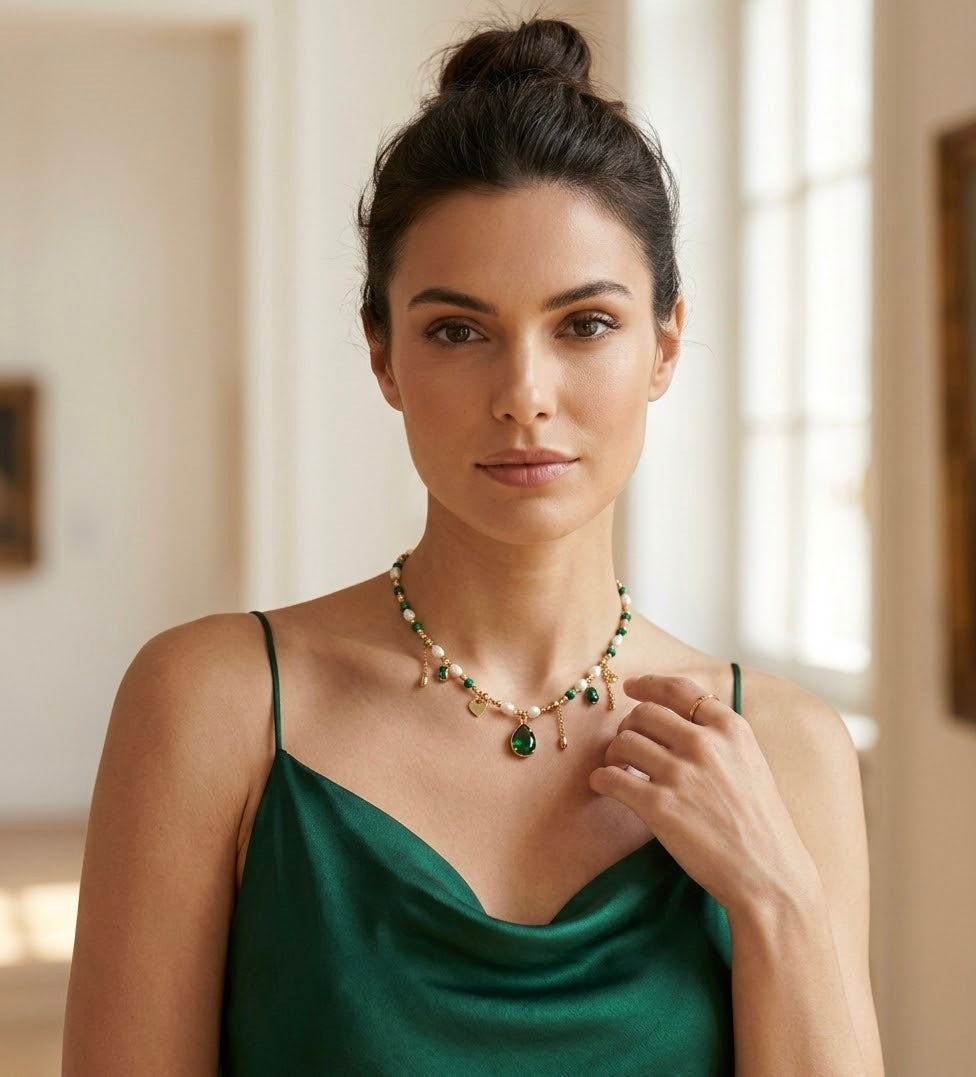 A woman with dark hair styled in a high bun wears a green satin cowl-neck camisole top and a beaded charm necklace. The necklace features pearls, green beads, gold accents, and a large teardrop-shaped green gemstone pendant that sits near the center of her collarbone.
